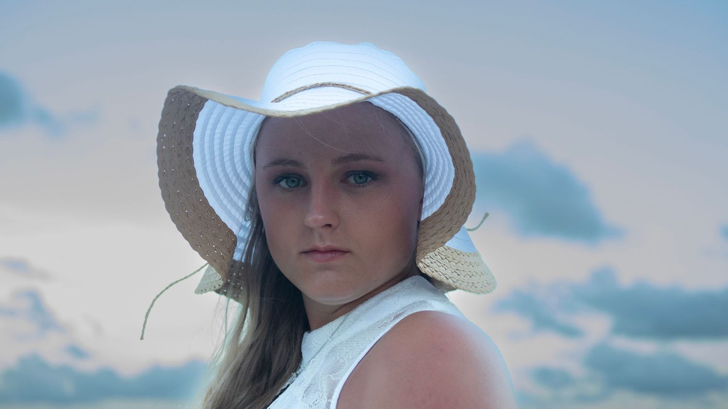 Portrait of someone in a white sun hat against a cloudy blue sky.