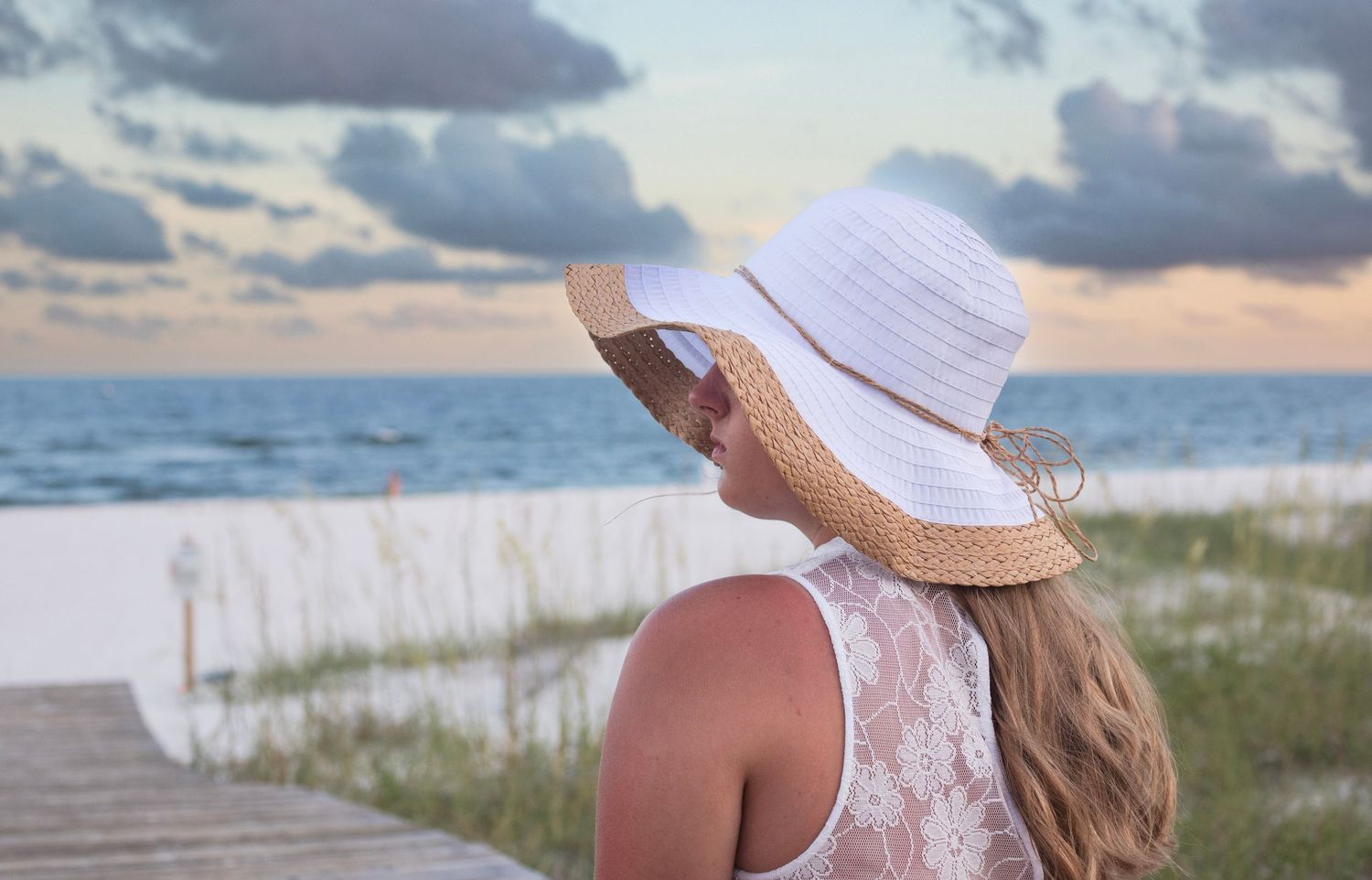 Person in white dress and sunhat sitting on boardwalk at beach during sunset.