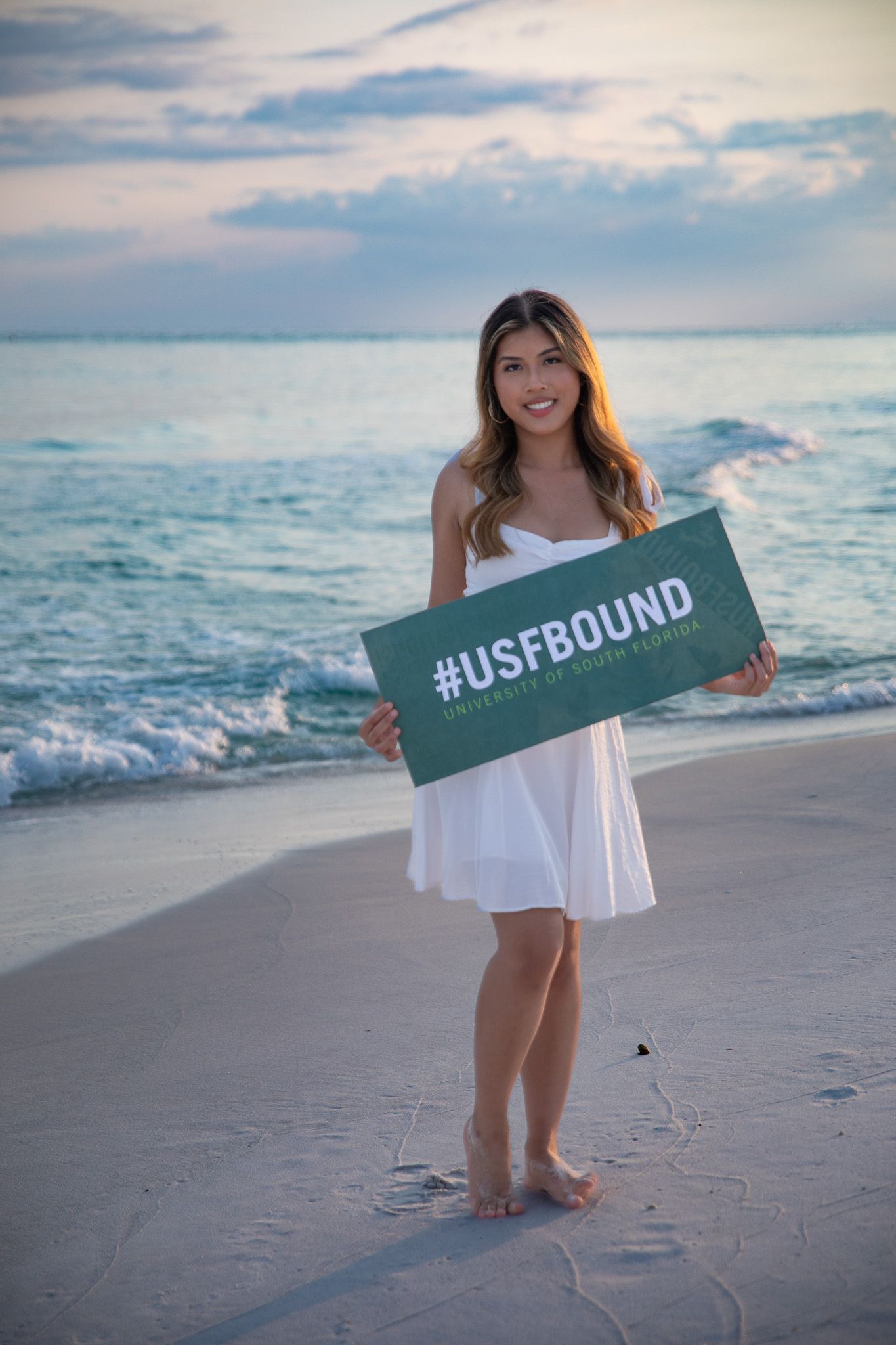Person in white dress holds green #USFBOUND sign while standing on beach at sunset.