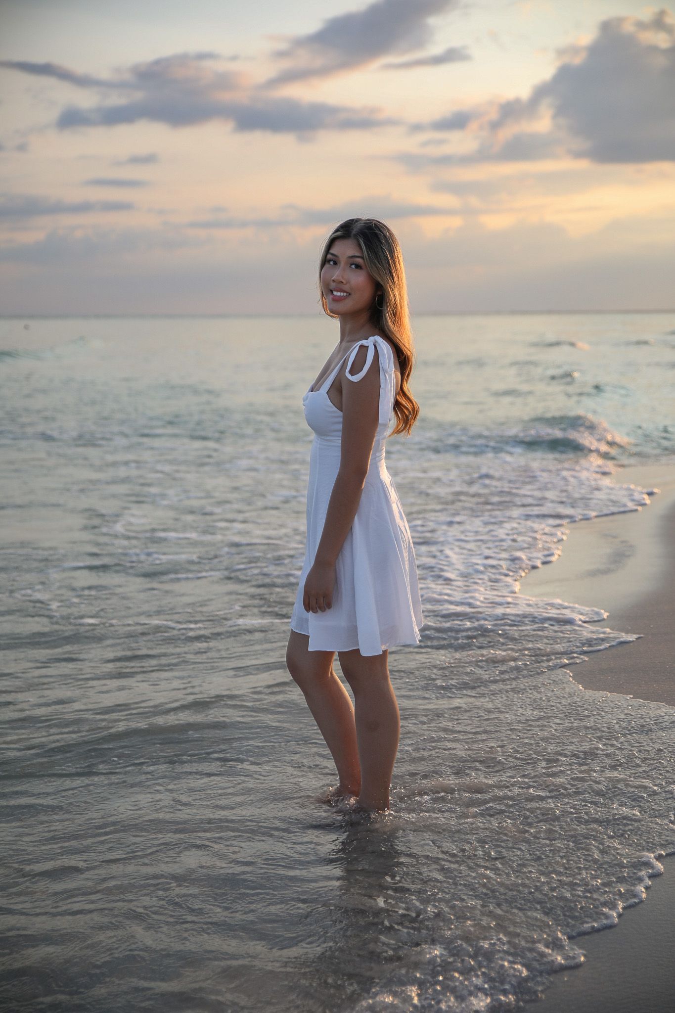 Woman in white sundress poses at sunset on a tropical beach with waves rolling in.