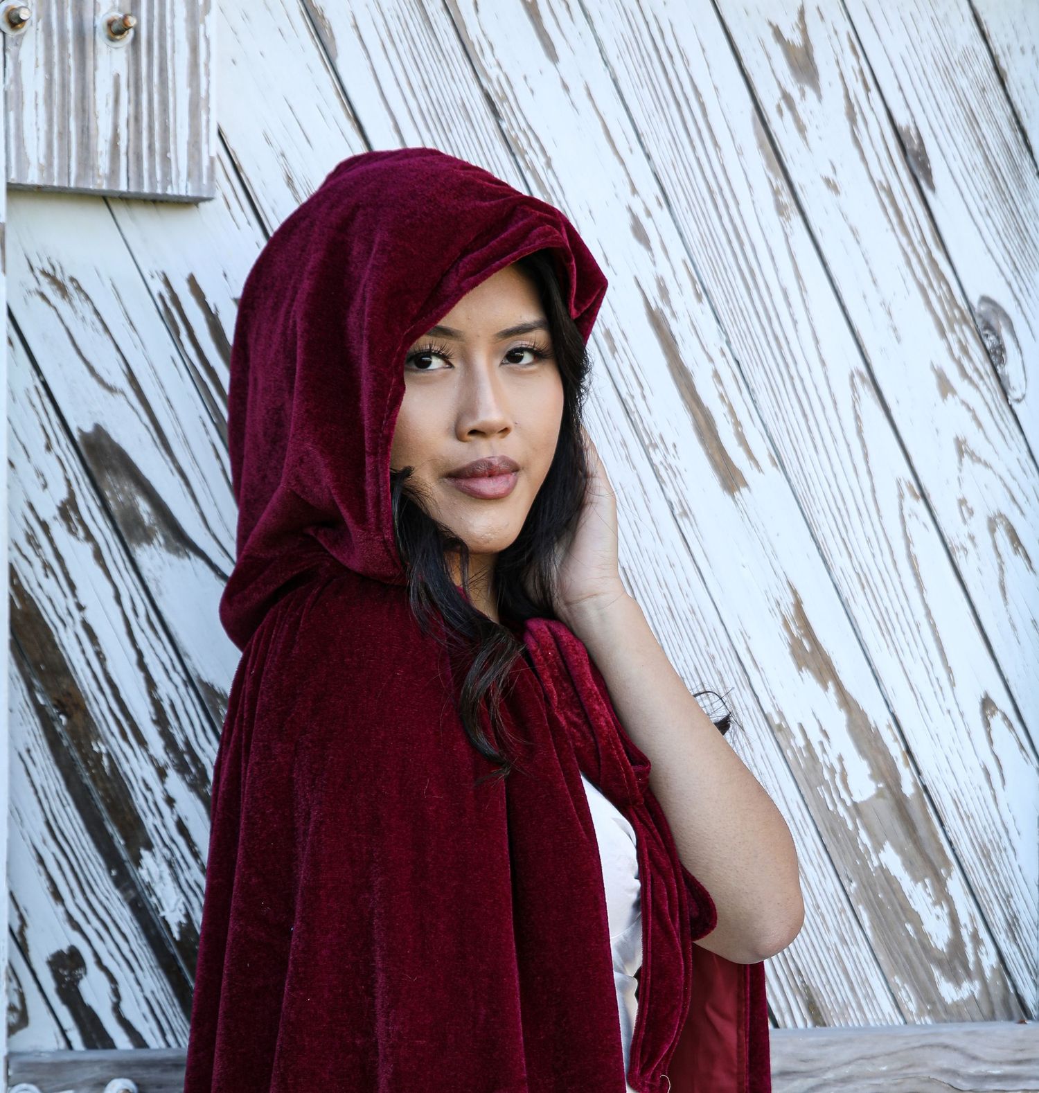 Portrait wearing deep red velvet hooded cape against white wooden backdrop.