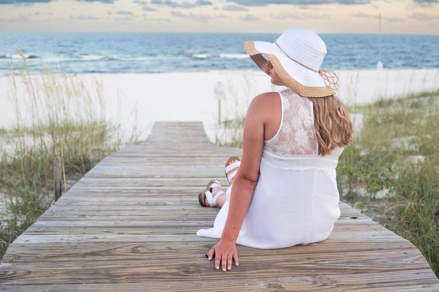 Someone in white dress and sunhat sits on wooden boardwalk overlooking ocean at sunset.