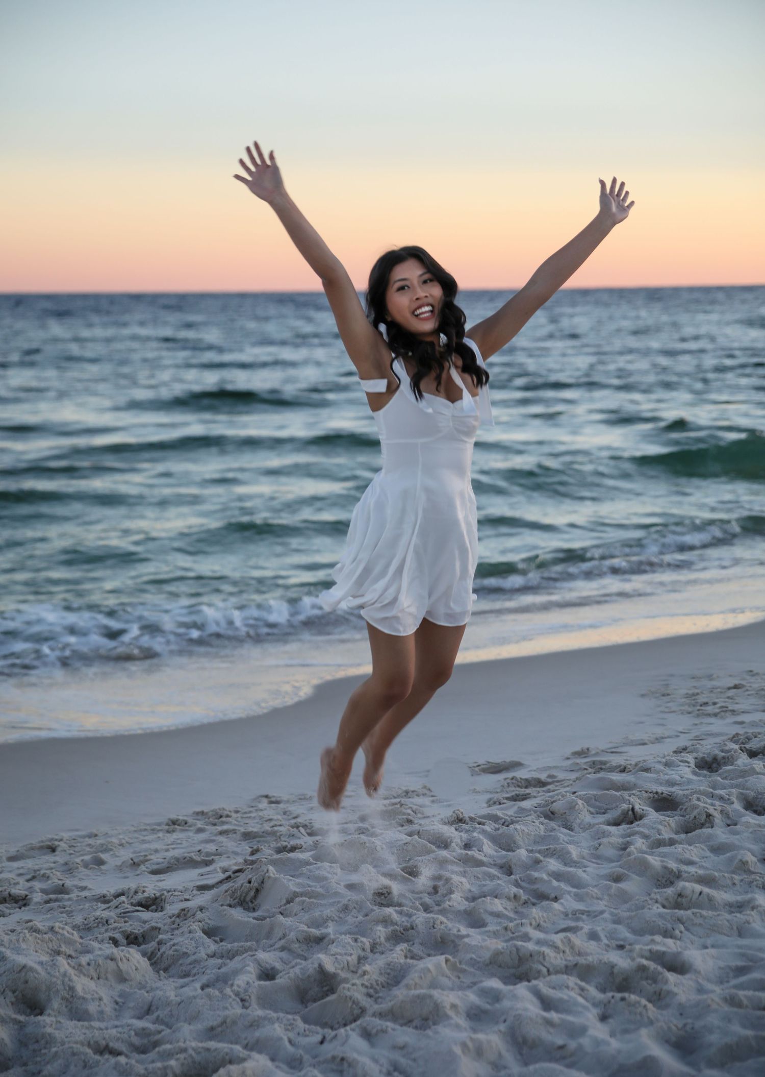 Person in white dress jumps joyfully on beach at sunset with arms raised.