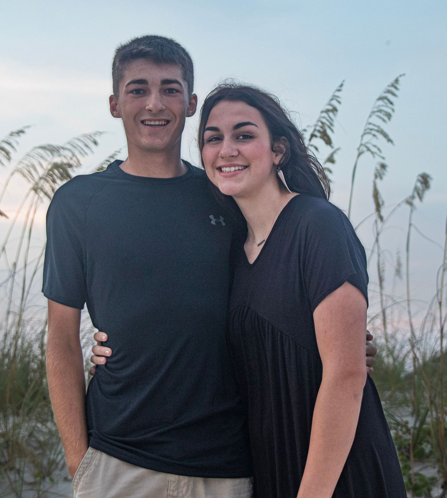 Two people in dark shirts pose together against coastal vegetation at dusk.