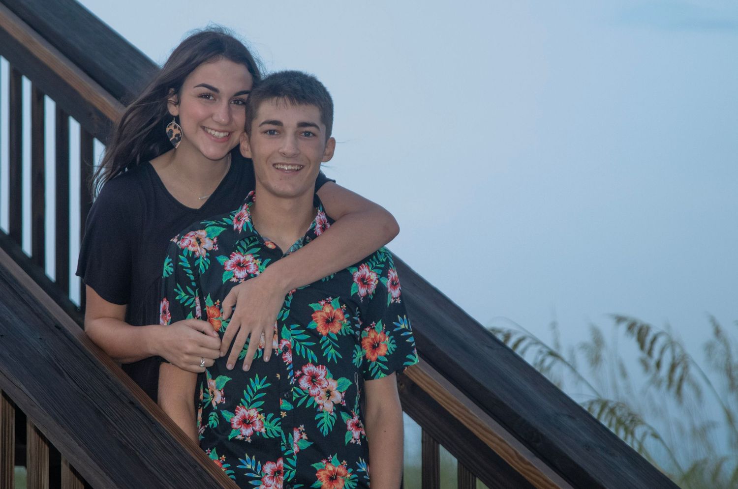 Two people pose together on outdoor stairs with coastal scenery in background.