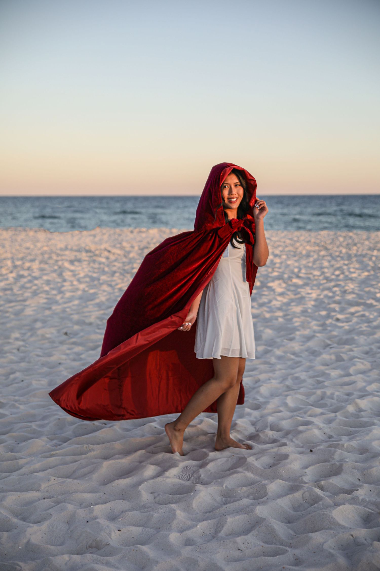 Figure in white dress and flowing red cape poses dramatically on a sandy beach at sunset.