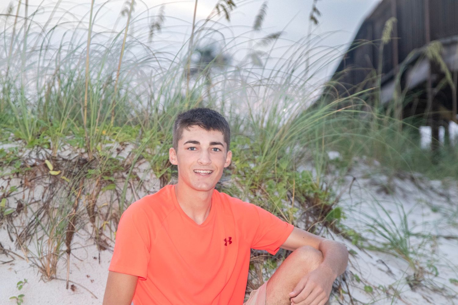 Person in coral orange shirt sits casually on beach sand with sea oats in background.