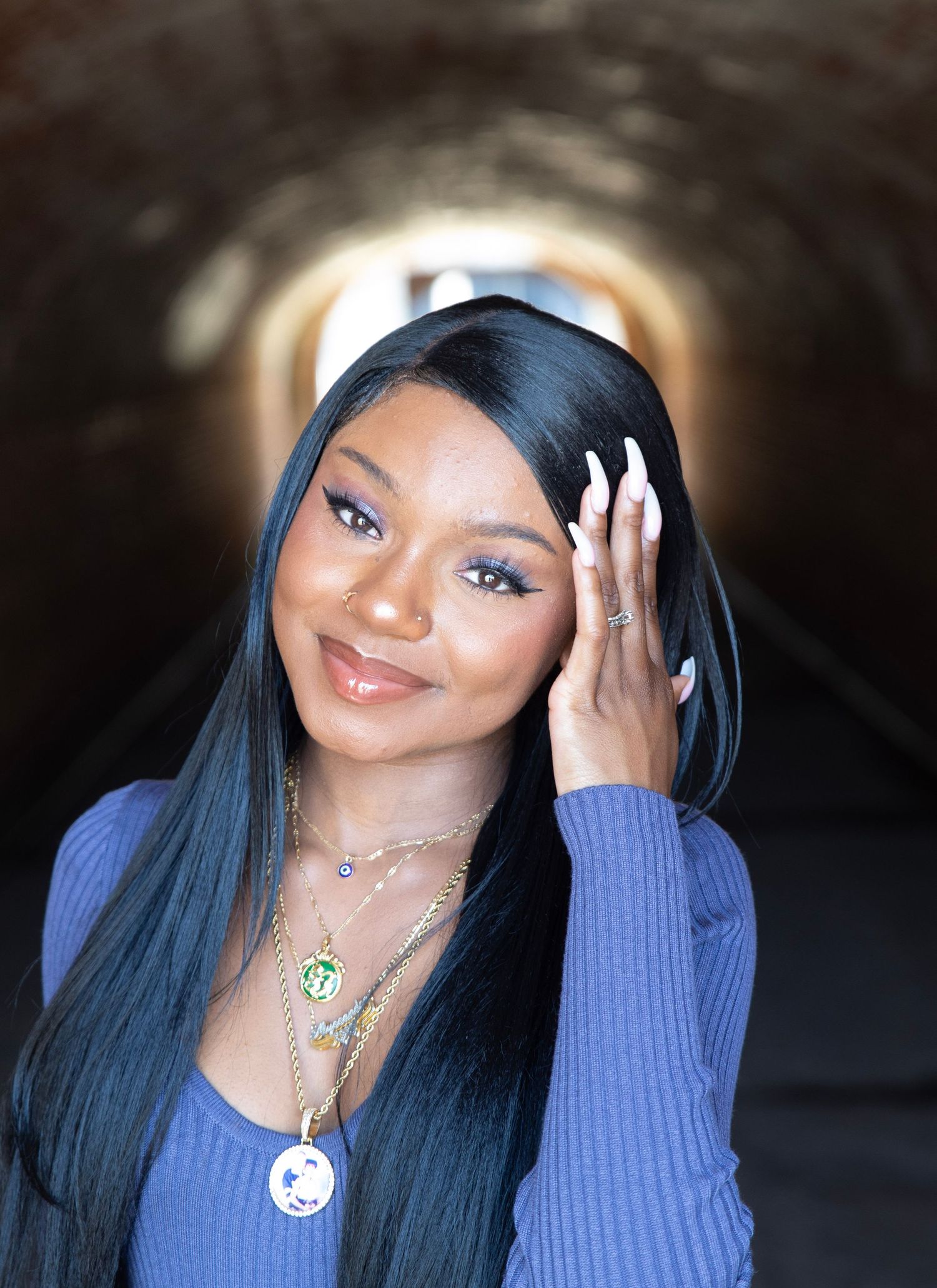 Portrait of person with long blue-black hair wearing purple sweater and necklaces in tunnel setting.