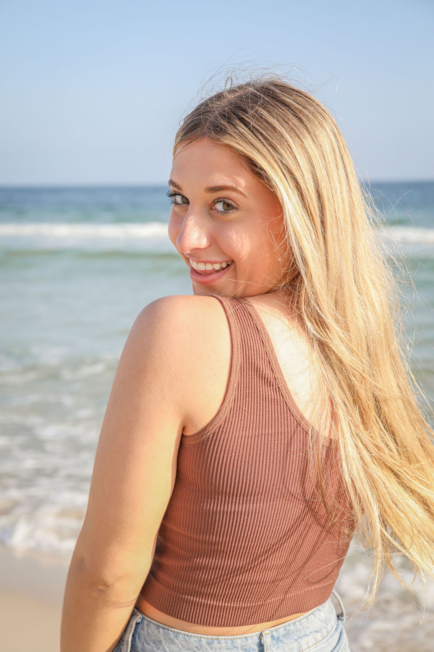 Sun-kissed beachgoer in brown tank top smiling over shoulder with ocean waves behind.