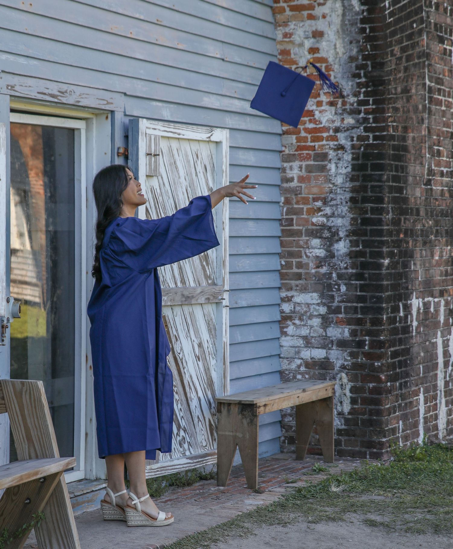 Graduate in blue cap and gown tosses cap in celebratory pose outside weathered building.