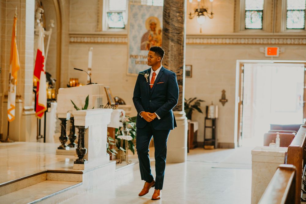 Handsome groom in navy suit and floral tie waits at the altar in a grand church.