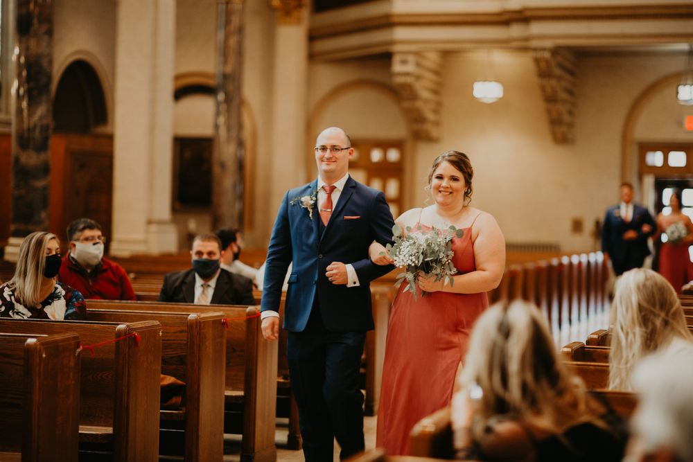 Bridesmaid in coral dress walks down church aisle with groomsman during wedding processional.