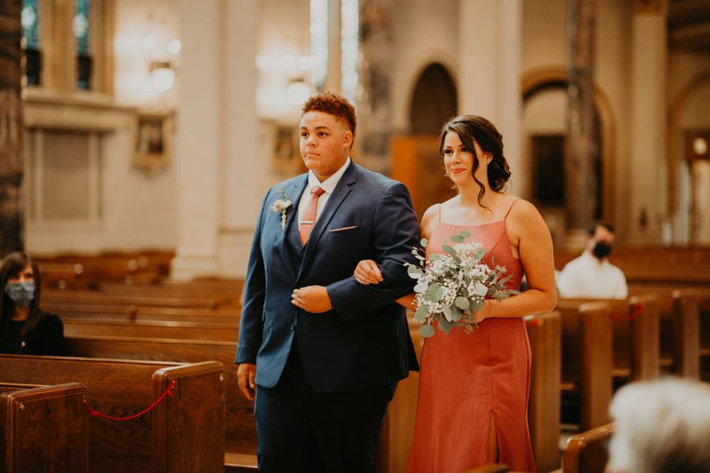 Groomsman in navy suit escorts bridesmaid in coral dress down the aisle at church wedding.