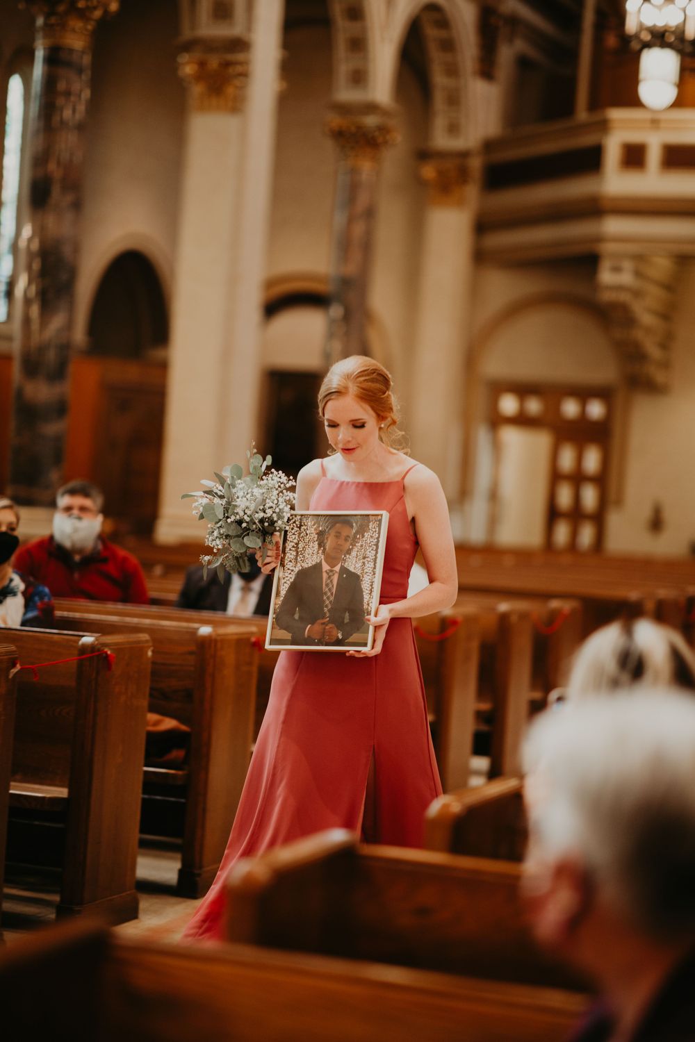Bridesmaid in pink dress carries framed photo and bouquet down church aisle at wedding ceremony.