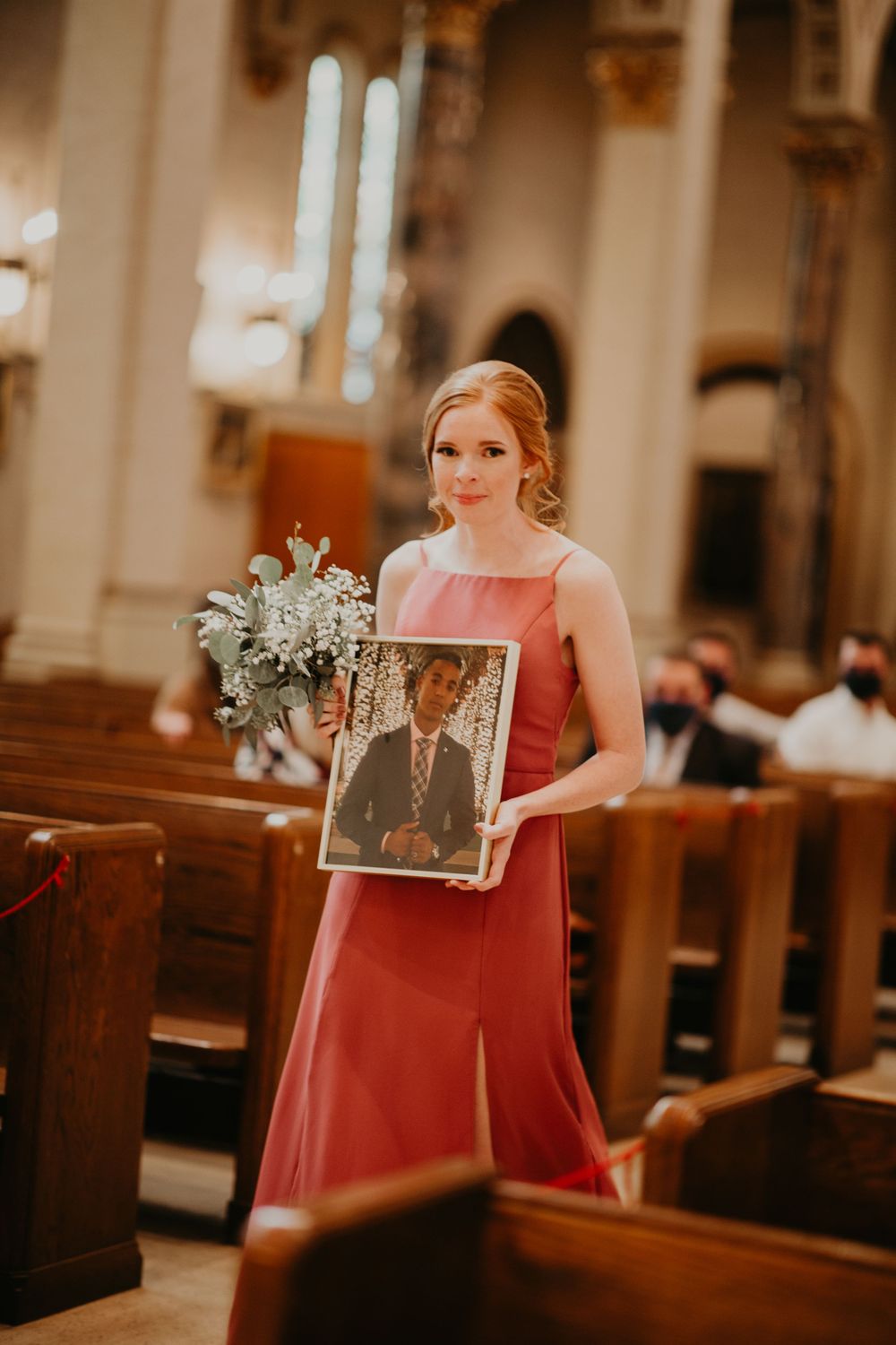 Bridesmaid in rose gown carries bouquet and memorial portrait down church aisle at wedding.