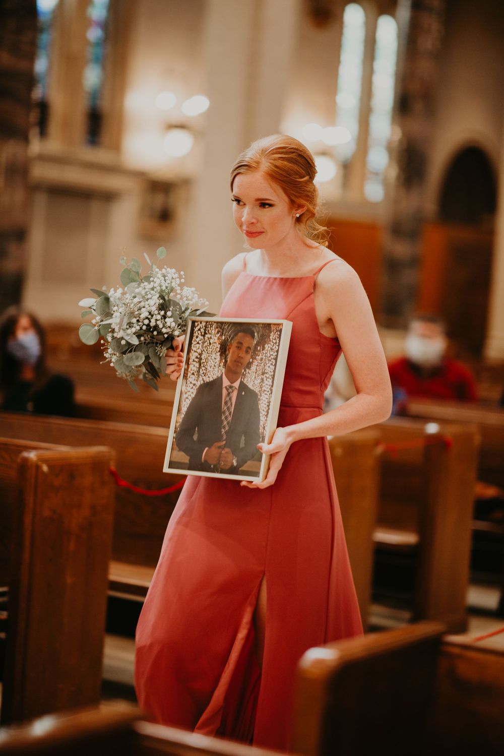 Bridesmaid in coral dress walks down church aisle holding bouquet and framed portrait at wedding.