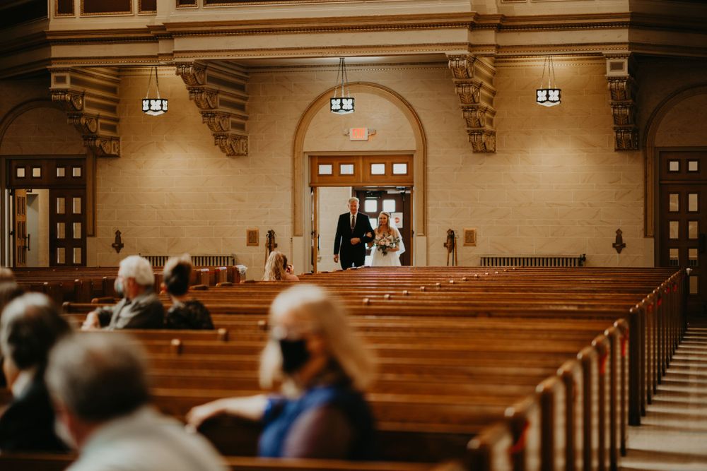 Bride walks down the aisle of an ornate church with wooden pews during wedding ceremony.