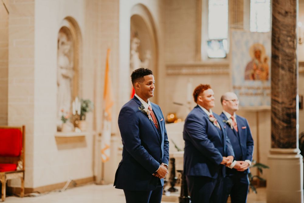 Groom and groomsmen in navy suits stand at church altar awaiting bride during wedding ceremony.