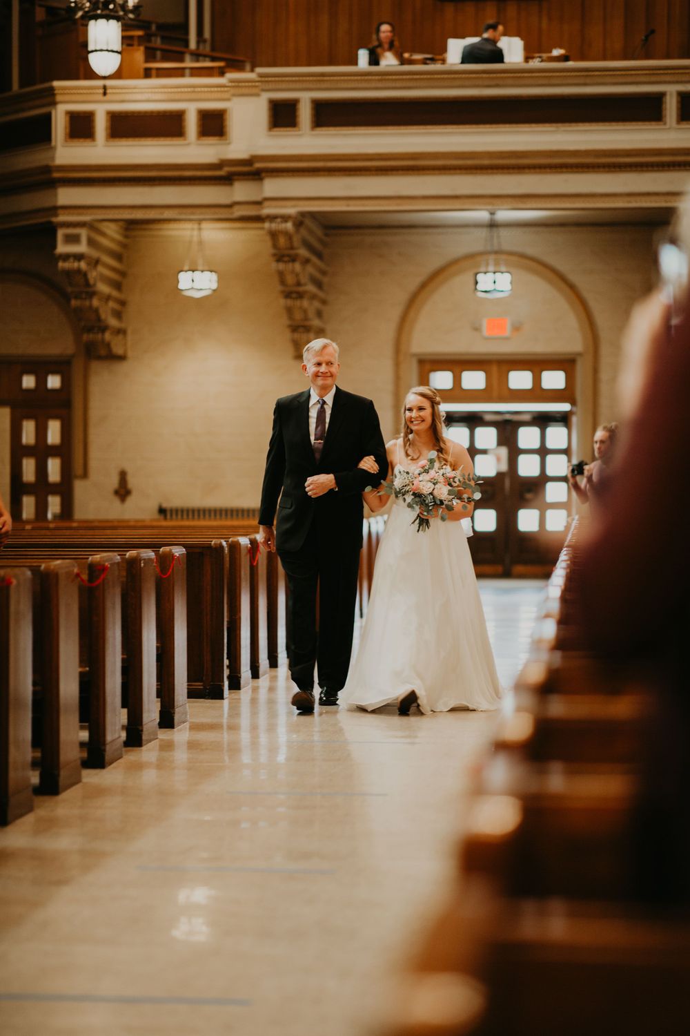 Smiling bride in white gown walks down church aisle with father during wedding processional.