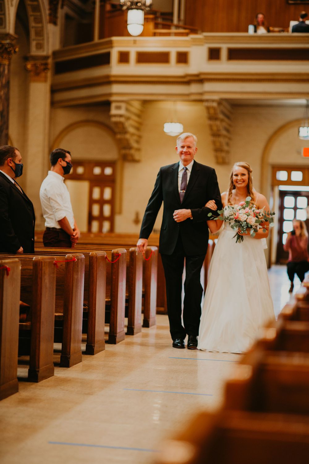 Bride and father walk down church aisle during wedding processional with guests watching nearby.