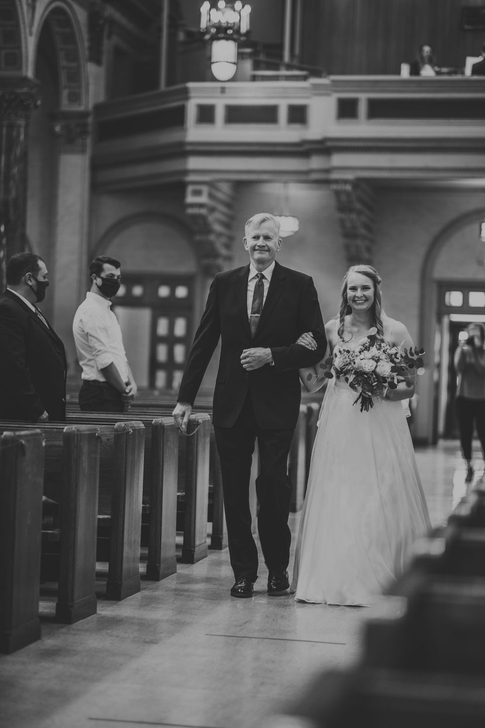 Black and white photo of bride and father walking down church aisle during wedding processional.