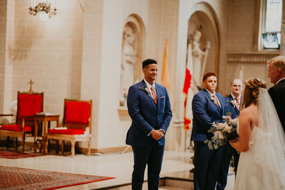 Emotional groom in navy suit smiles seeing bride approach altar during church wedding ceremony.