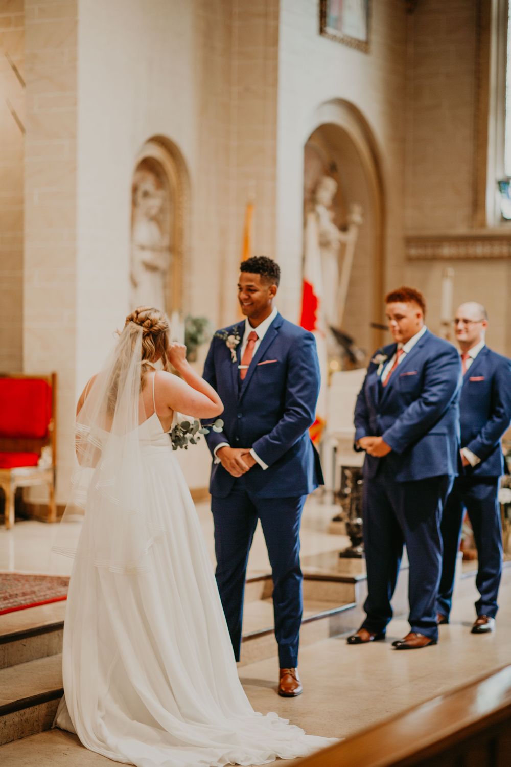 Groom in blue suit smiles at bride in white gown during church wedding ceremony at the altar.