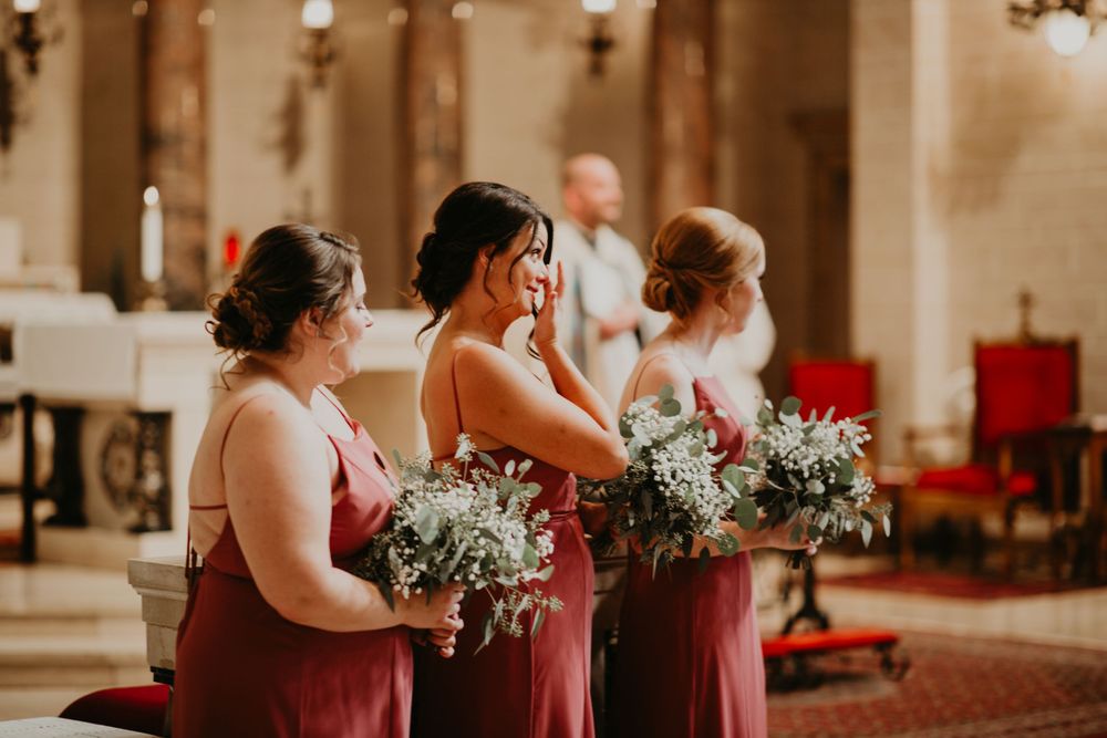 Bridesmaids in dusty rose gowns holding greenery bouquets wipe tears during emotional church wedding.