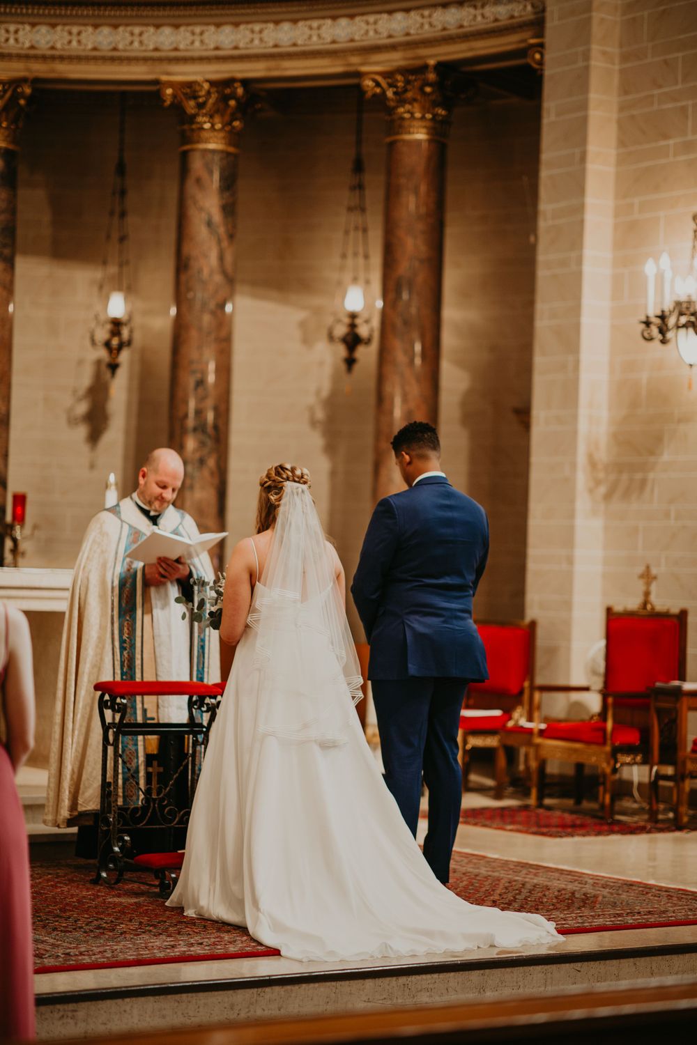 Smiling priest reads from book as bride and groom stand at altar during church wedding ceremony.