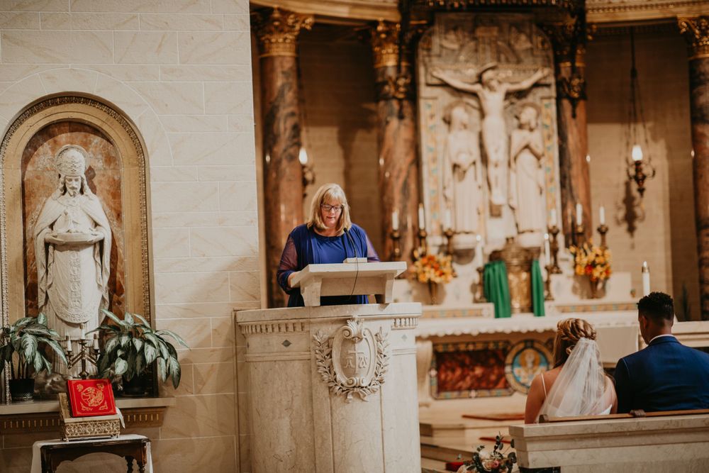 Female reader at ornate church pulpit delivers reading during wedding ceremony beside Virgin Mary statue.