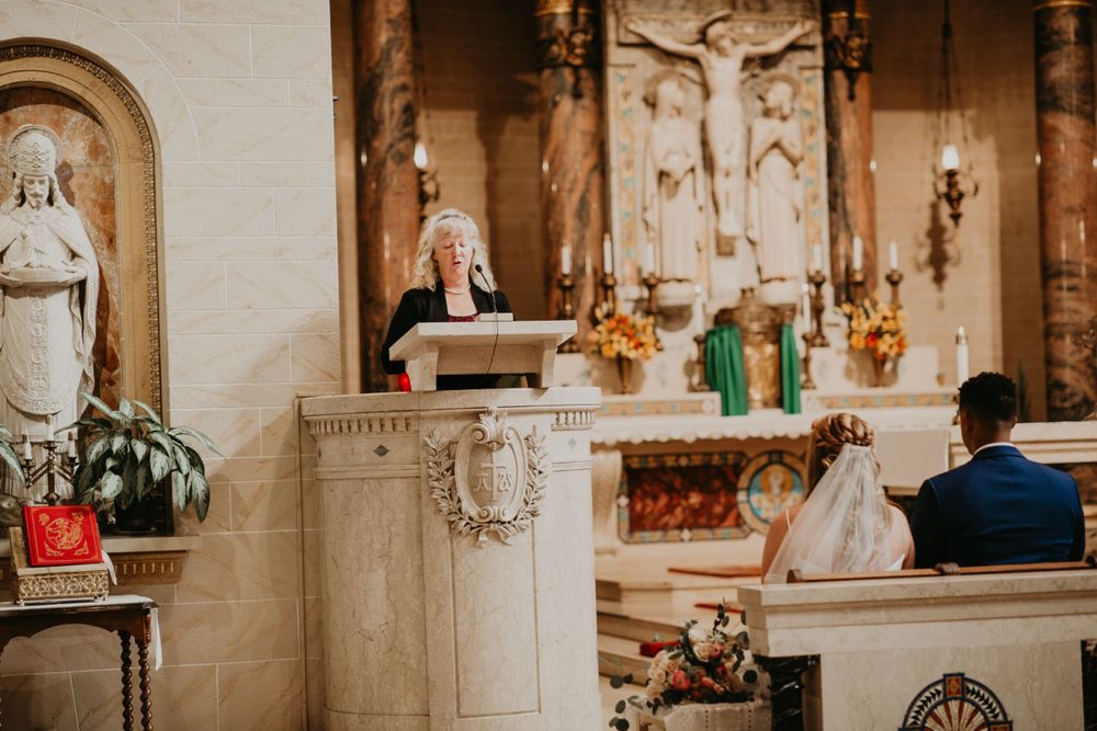 Woman in blue reads scripture at church podium while bride and groom sit during wedding ceremony.
