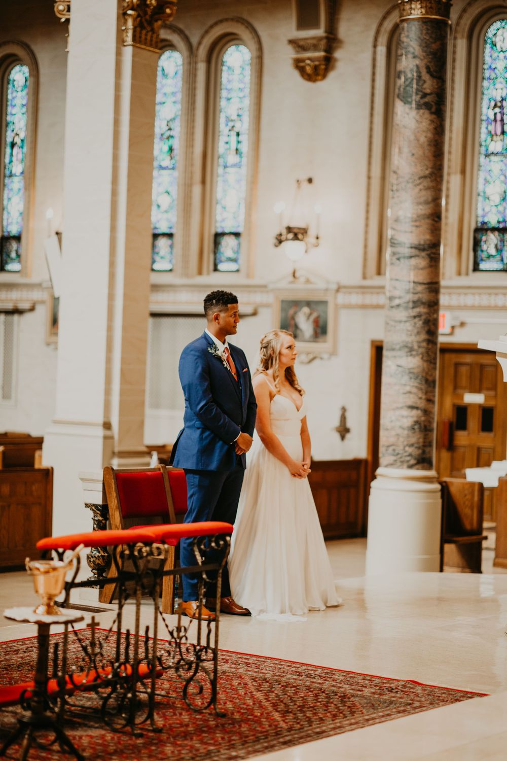 Bride in white gown and groom in blue suit stand together in sunlit church during wedding ceremony.