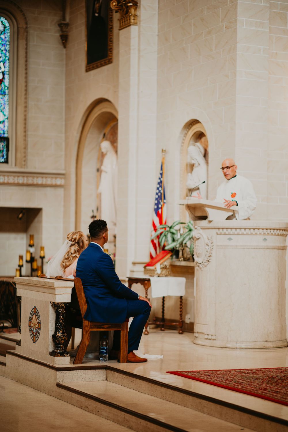Groom in blue suit seated at altar while officiant delivers wedding sermon at pulpit.