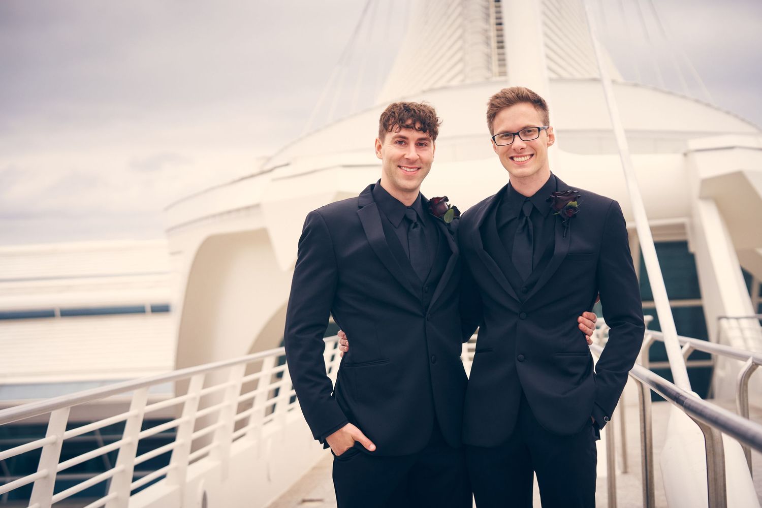 Two people in black suits pose together on a cruise ship deck with white railings visible.