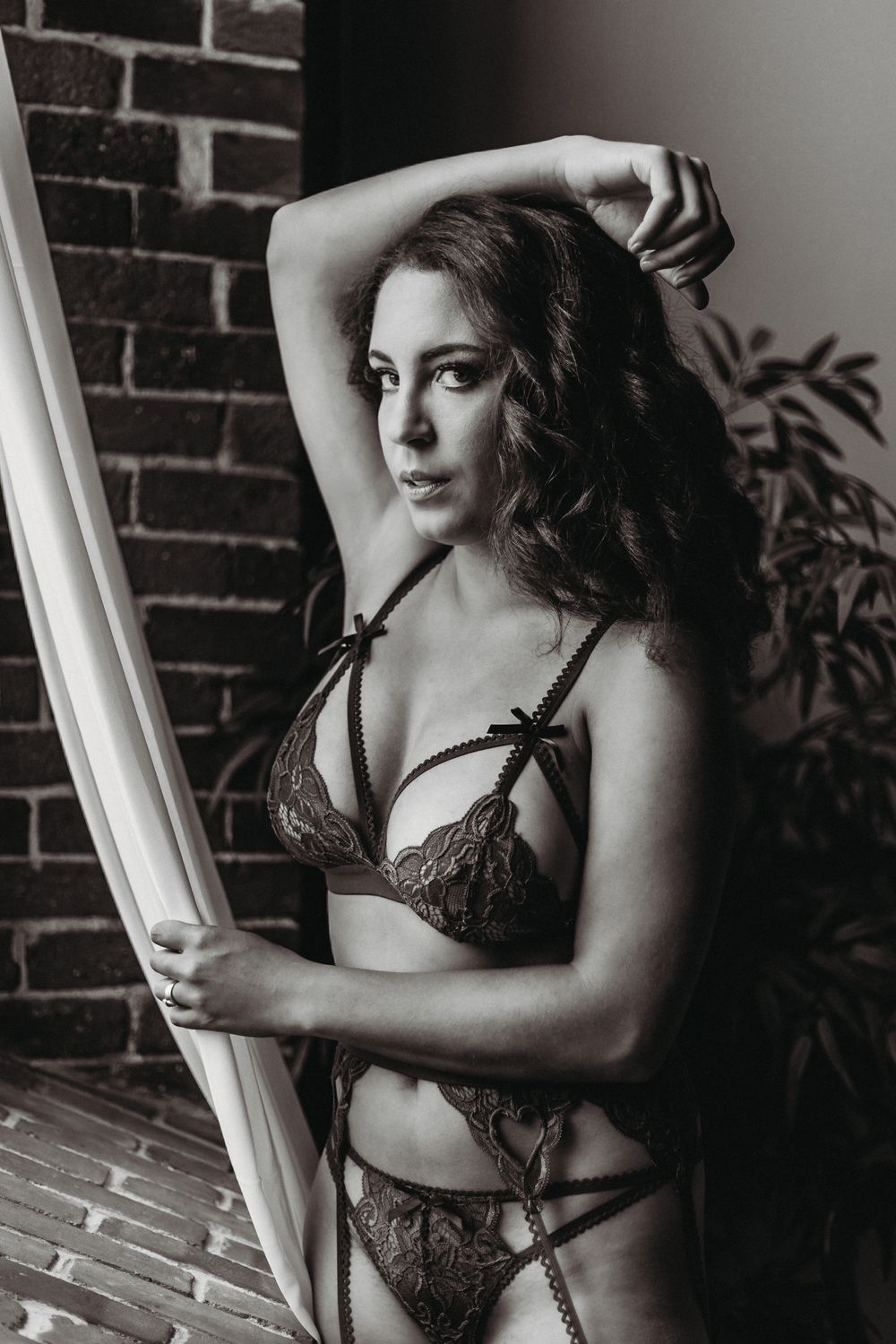 Black and white artistic boudoir photo of a woman in lace lingerie posing against a brick wall.