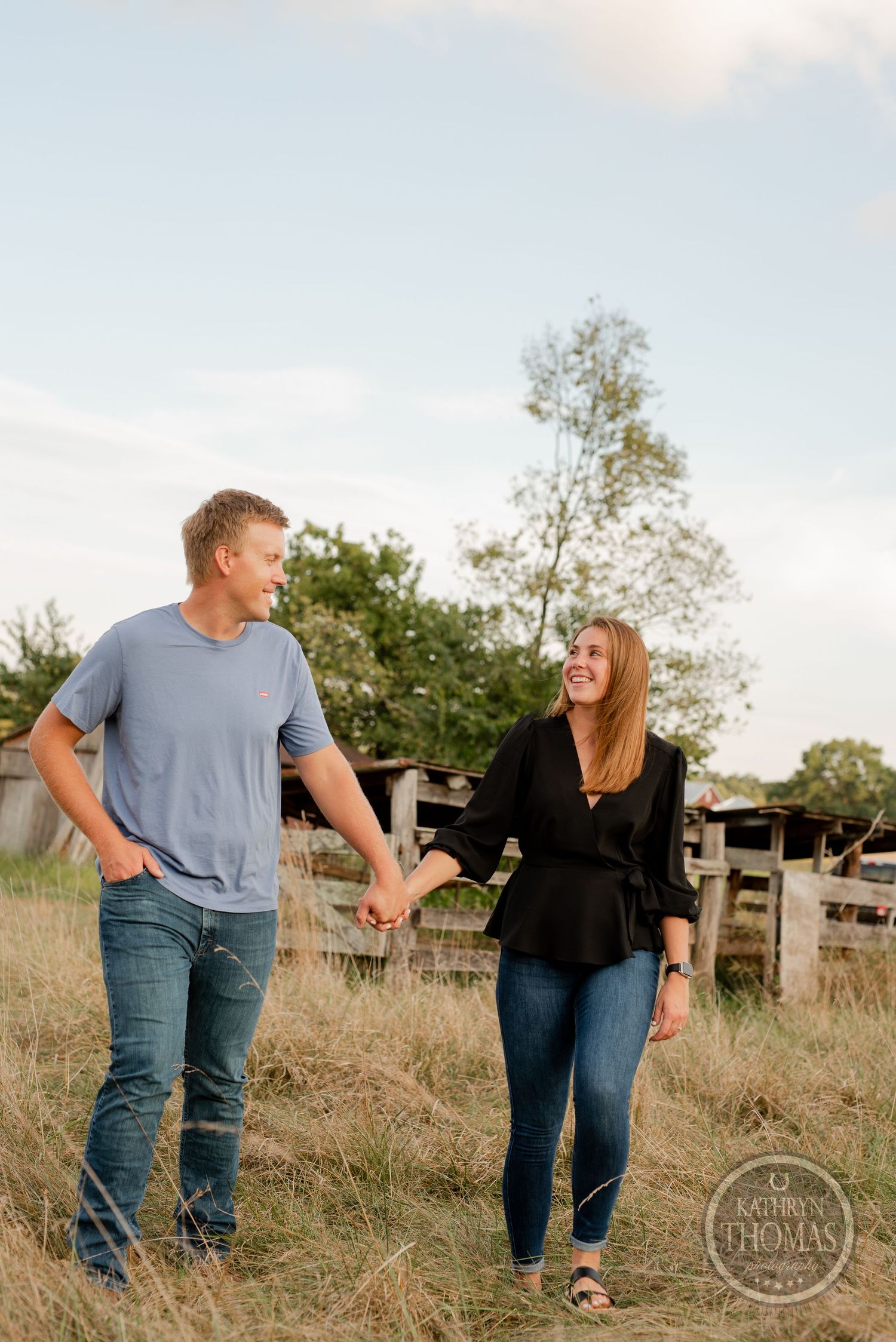Couple holding hands while walking in a grassy field with wooden fence in background.