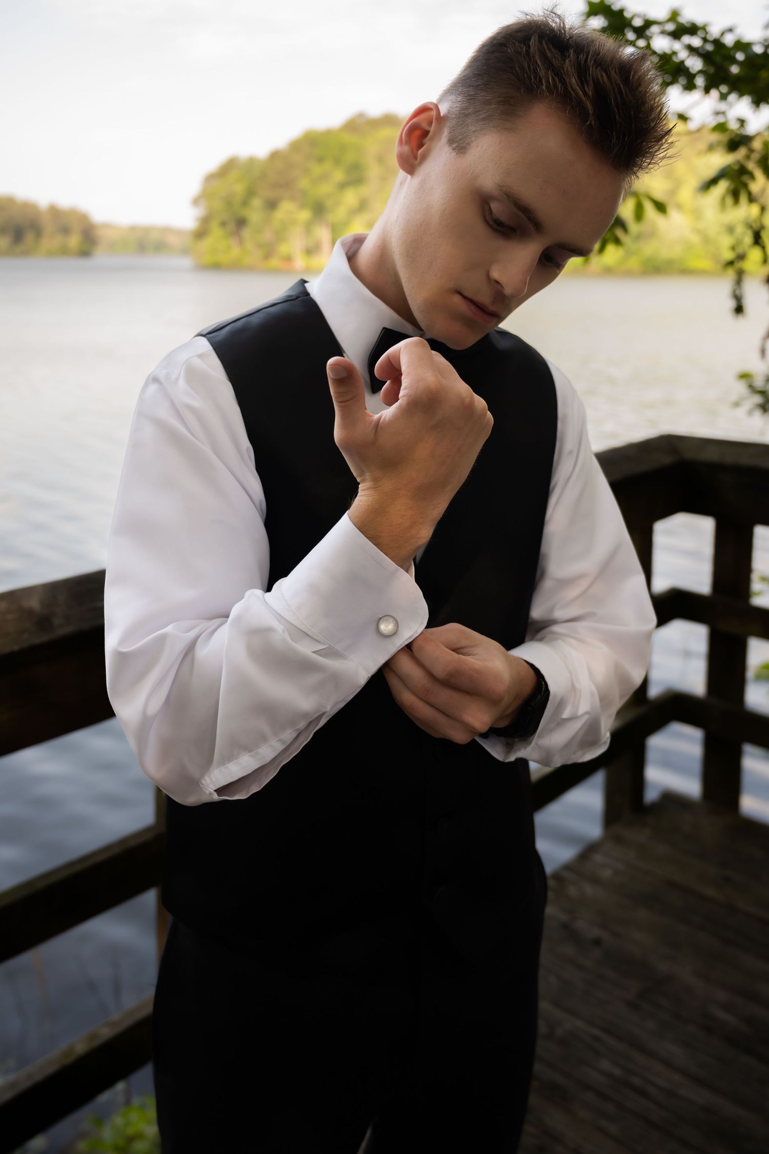 A figure wearing formal attire adjusts their cufflinks on a lakeside deck.