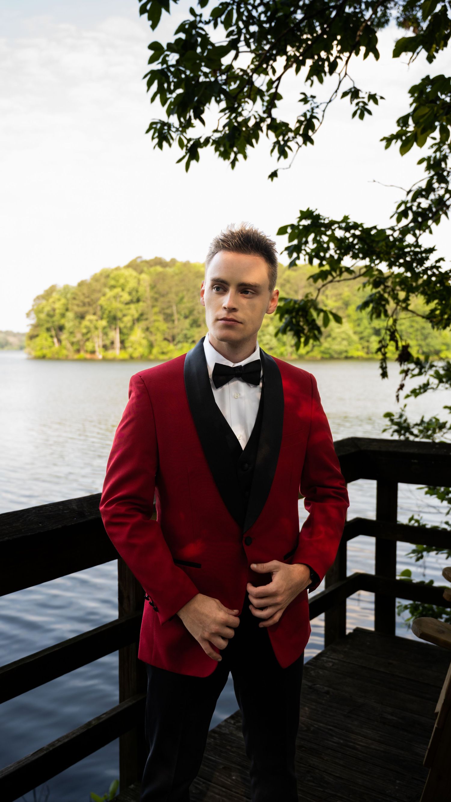 A person in a red formal jacket and black bowtie poses near a waterfront wooden deck.