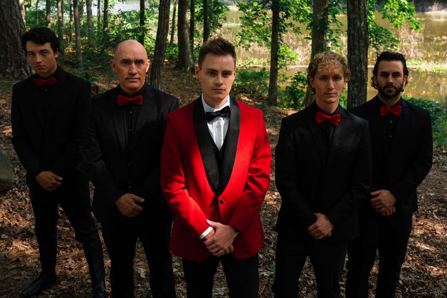 Groomsmen in black and red formal attire pose together in a wooded setting.