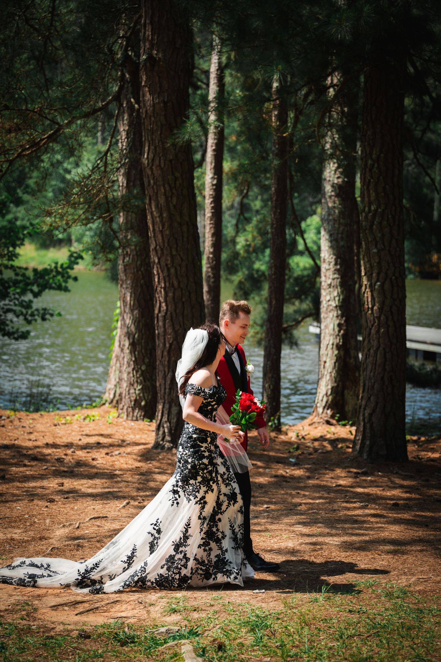 A couple shares an intimate moment among pine trees near a lake, wearing formal black and white attire.