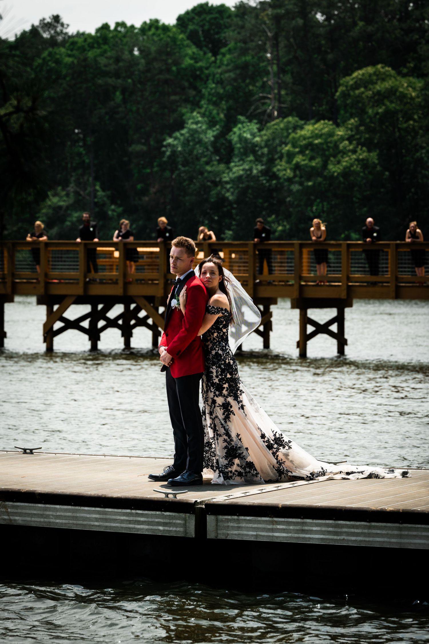 A couple in formal wear poses on a dock with onlookers visible on a bridge in the background.