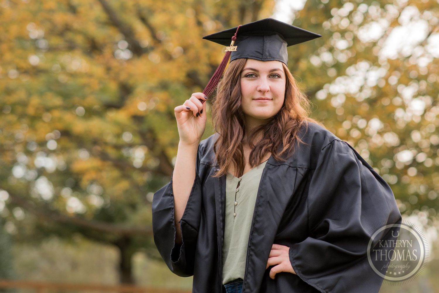 A graduate in cap and gown poses outdoors during autumn with trees in background.