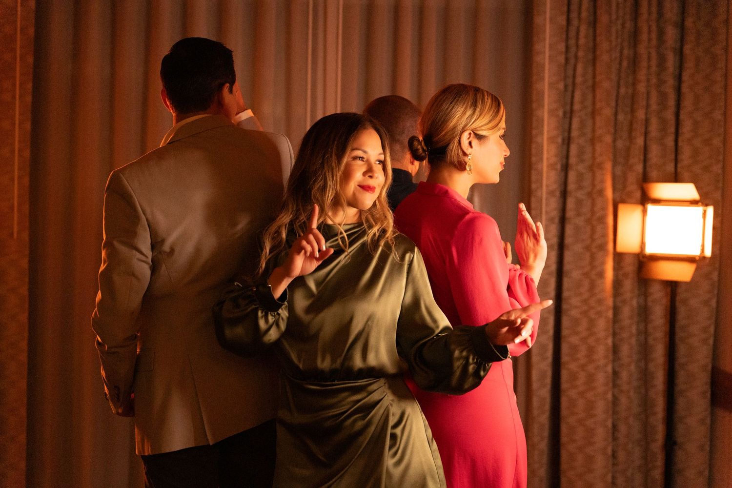 Three people stand close together in formal attire at an evening event with warm lighting and curtains in the background.