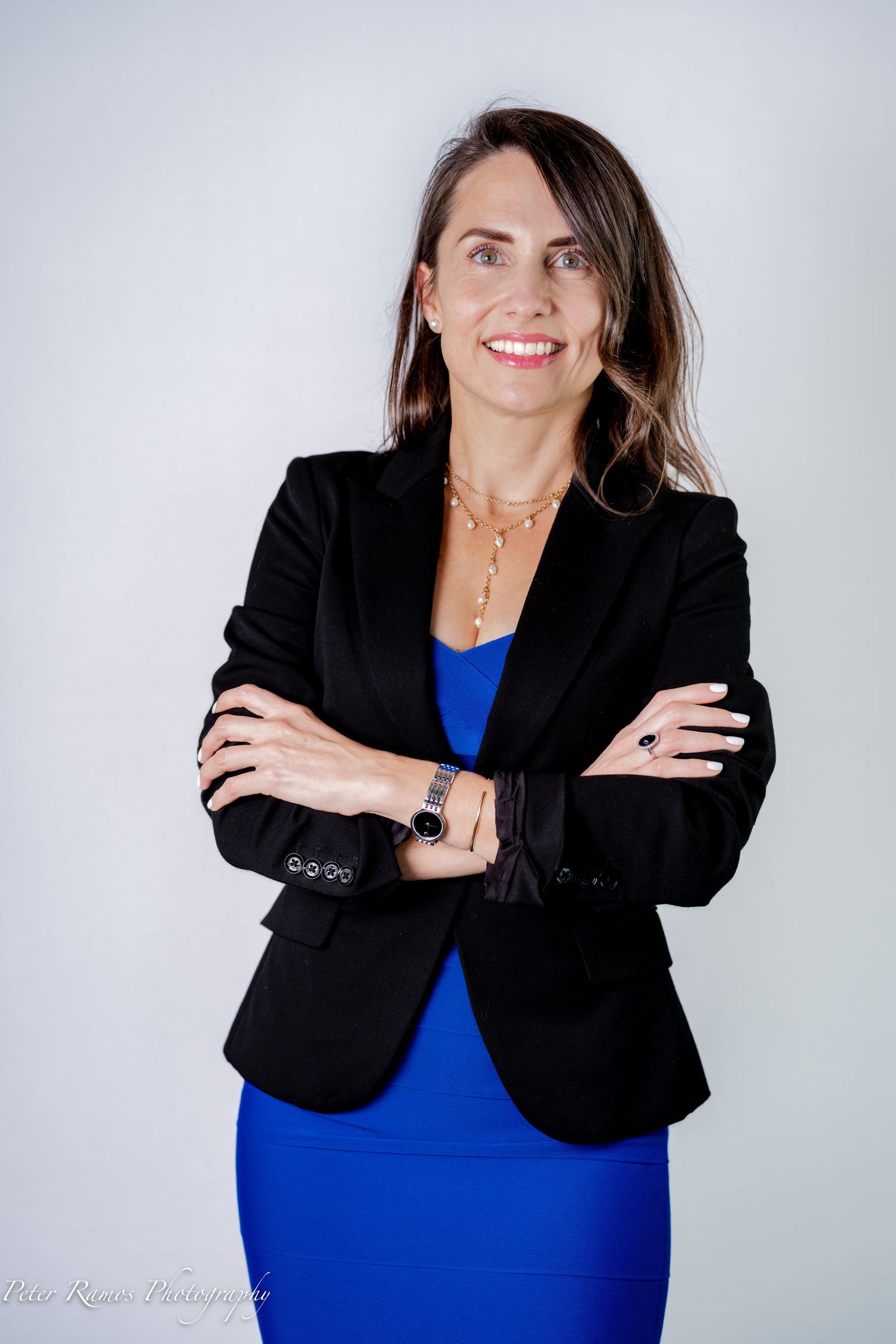 Professional portrait of someone in a black blazer and blue dress with arms crossed and a confident smile.