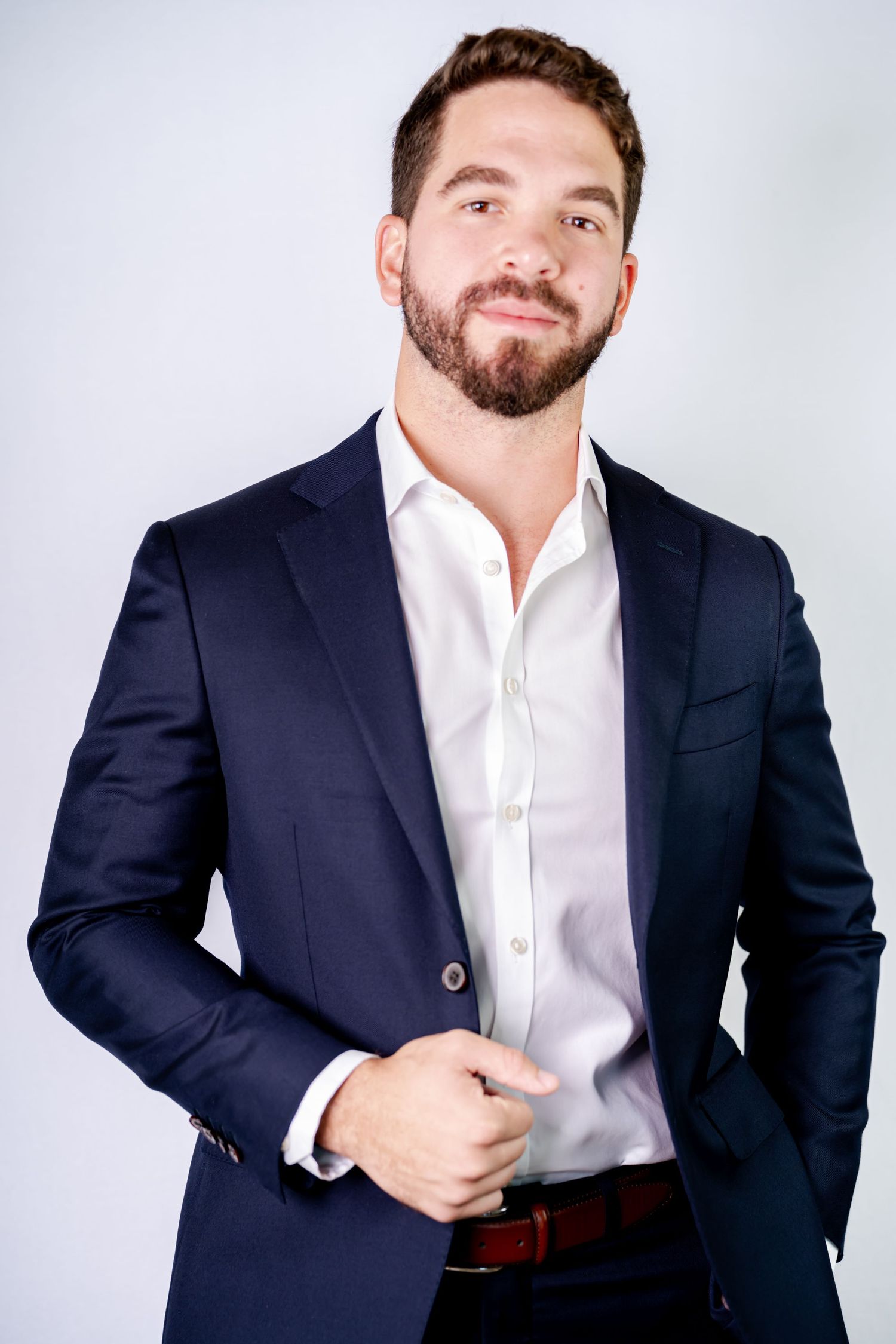 Professional headshot of a bearded person wearing a navy suit jacket with white dress shirt against light background.