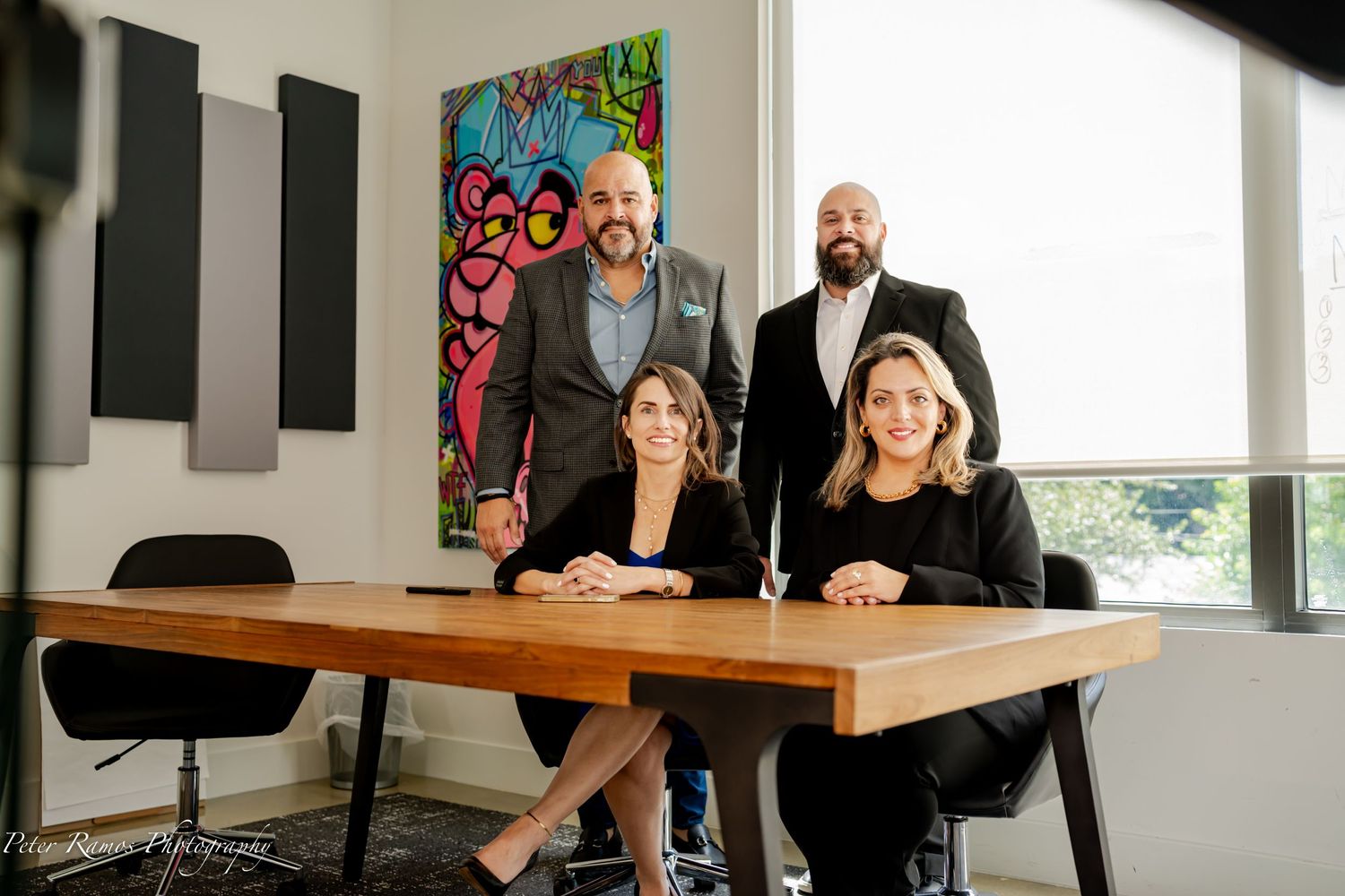 Four business professionals sit and stand around a wooden conference table in a modern office with colorful wall art.