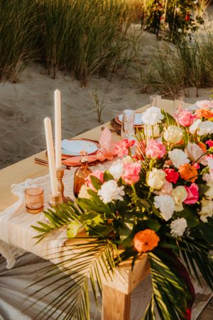 A romantic beach wedding table setup with coral and white flowers, candles, and palm fronds at sunset.