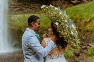 Newlyweds share an intimate moment under a floral umbrella by a waterfall.