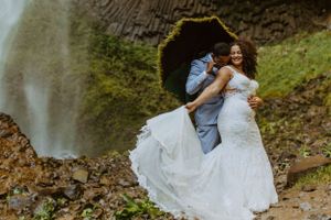 Couple in wedding attire dancing near a scenic waterfall.