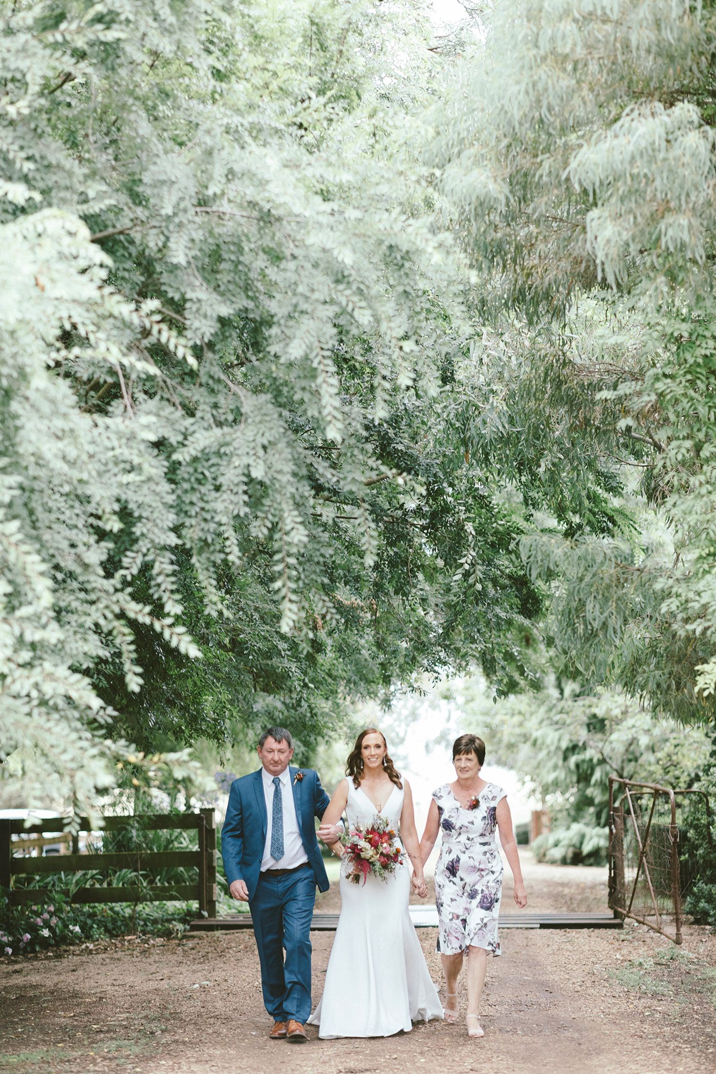 Wedding party walks down tree-lined path at outdoor venue.